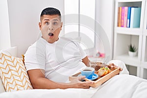 Young hispanic man eating breakfast in the bed scared and amazed with open mouth for surprise, disbelief face