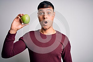 Young hispanic man eating fresh green apple as healthy nutrition over isolated background scared in shock with a surprise face,