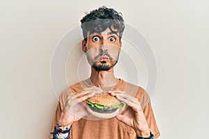 Young hispanic man eating a tasty classic burger puffing cheeks with funny face