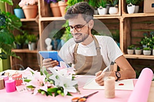 Young hispanic man florist using touchpad writing on notebook at flower shop