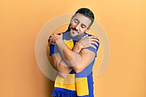 Young hispanic man football hooligan cheering game hugging oneself happy and positive, smiling confident