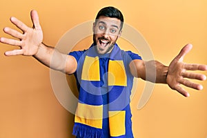 Young hispanic man football hooligan cheering game looking at the camera smiling with open arms for hug