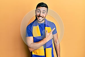 Young hispanic man football hooligan cheering game smiling cheerful pointing with hand and finger up to the side