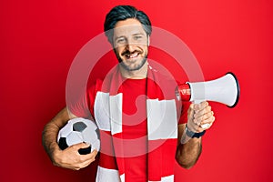 Young hispanic man football hooligan holding ball using megaphone smiling with a happy and cool smile on face