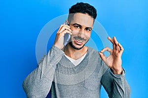 Young hispanic man having conversation talking on the smartphone doing ok sign with fingers, smiling friendly gesturing excellent