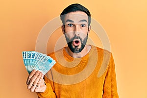 Young hispanic man holding 100 brazilian real banknotes scared and amazed with open mouth for surprise, disbelief face