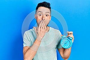 Young hispanic man holding alarm clock covering mouth with hand, shocked and afraid for mistake