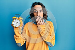 Young hispanic man holding alarm clock covering mouth with hand, shocked and afraid for mistake