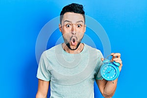 Young hispanic man holding alarm clock scared and amazed with open mouth for surprise, disbelief face