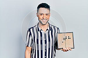 Young hispanic man holding asian go game board looking positive and happy standing and smiling with a confident smile showing