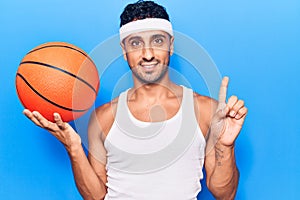 Young hispanic man holding basketball ball smiling with an idea or question pointing finger with happy face, number one