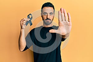 Young hispanic man holding black ribbon with open hand doing stop sign with serious and confident expression, defense gesture