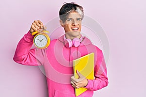 Young hispanic man holding book and alarm clock clueless and confused expression