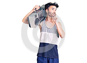 Young hispanic man holding boombox, listening to music serious face thinking about question with hand on chin, thoughtful about