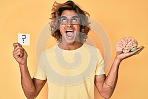 Young hispanic man holding brain and question mark angry and mad screaming frustrated and furious, shouting with anger looking up