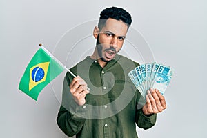 Young hispanic man holding brazil flag and real banknotes in shock face, looking skeptical and sarcastic, surprised with open