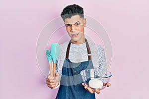 Young hispanic man holding bread dough and cooking tools in shock face, looking skeptical and sarcastic, surprised with open mouth