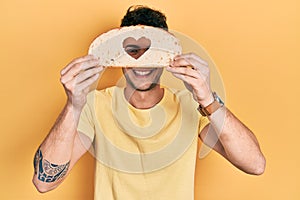 Young hispanic man holding bread with heart shape over eye smiling and laughing hard out loud because funny crazy joke