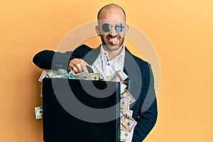 Young hispanic man holding briefcase with dollars sticking tongue out happy with funny expression