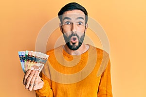 Young hispanic man holding canadian dollars scared and amazed with open mouth for surprise, disbelief face