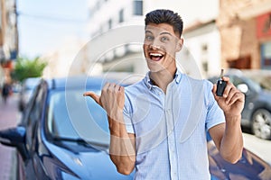 Young hispanic man holding car key pointing thumb up to the side smiling happy with open mouth
