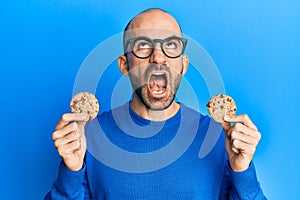 Young hispanic man holding chocolate chips cookies angry and mad screaming frustrated and furious, shouting with anger looking up