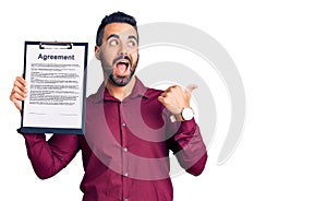 Young hispanic man holding clipboard with agreement document pointing thumb up to the side smiling happy with open mouth