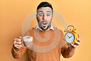 Young hispanic man holding coffee and alarm clock afraid and shocked with surprise and amazed expression, fear and excited face