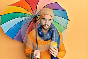 Young hispanic man holding colorful umbrella drinking take away coffee skeptic and nervous, frowning upset because of problem