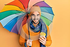 Young hispanic man holding colorful umbrella drinking take away coffee smiling with a happy and cool smile on face