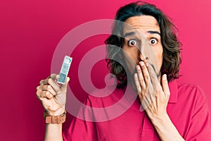 Young hispanic man holding computer memory covering mouth with hand, shocked and afraid for mistake