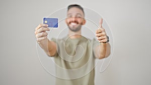 Young hispanic man holding credit card doing thumb up gesture smiling over isolated white background