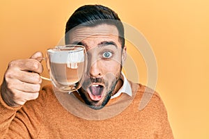 Young hispanic man holding cup of coffee over eye scared and amazed with open mouth for surprise, disbelief face