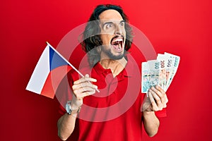 Young hispanic man holding czech republic flag and koruna banknotes angry and mad screaming frustrated and furious, shouting with