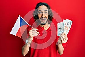 Young hispanic man holding czech republic flag and koruna banknotes celebrating crazy and amazed for success with open eyes