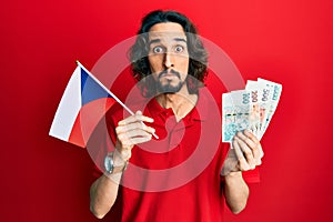 Young hispanic man holding czech republic flag and koruna banknotes puffing cheeks with funny face