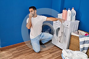 Young hispanic man holding dirty clothe closing nose at laundry room