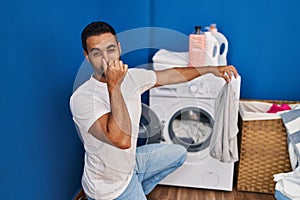 Young hispanic man holding dirty clothe closing nose at laundry room