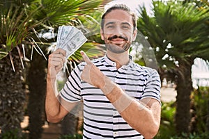 Young hispanic man holding dollars close to face smiling happy pointing with hand and finger