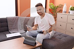 Young hispanic man holding dollars using calculator accounting at home