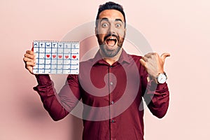 Young hispanic man holding heart calendar pointing thumb up to the side smiling happy with open mouth