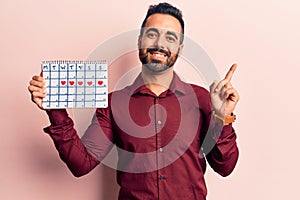 Young hispanic man holding heart calendar smiling happy pointing with hand and finger to the side