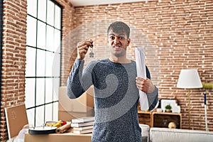 Young hispanic man holding keys of new home and blueprints clueless and confused expression