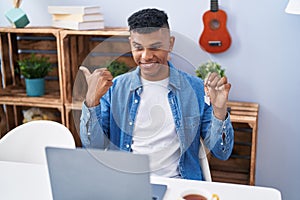 Young hispanic man holding keys of new home doing video call pointing thumb up to the side smiling happy with open mouth