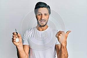 Young hispanic man holding keys of new home pointing thumb up to the side smiling happy with open mouth