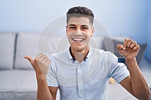 Young hispanic man holding keys of new home pointing thumb up to the side smiling happy with open mouth