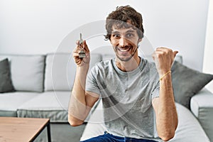 Young hispanic man holding keys of new home sitting on the sofa pointing thumb up to the side smiling happy with open mouth