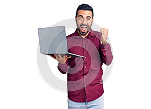 Young hispanic man holding laptop screaming proud, celebrating victory and success very excited with raised arms
