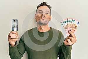 Young hispanic man holding led bulb and euros banknotes looking at the camera blowing a kiss being lovely and sexy