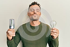Young hispanic man holding lightbulb for inspiration and idea looking at the camera blowing a kiss being lovely and sexy